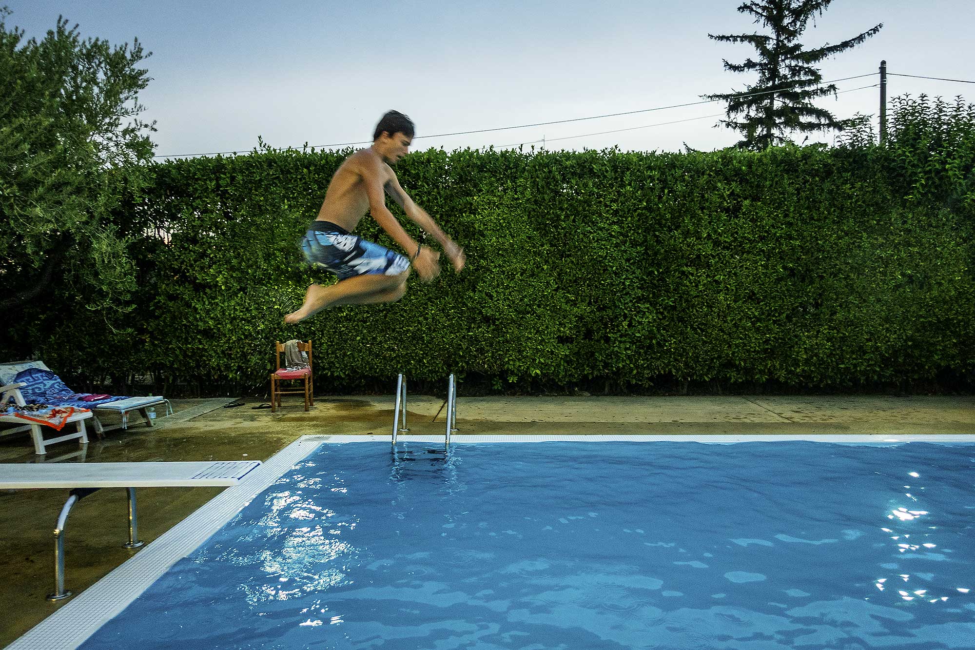 Teenage boy jumping into a backyard swimming pool, with diving board, pool ladder, and green hedge in the background on a summer day.