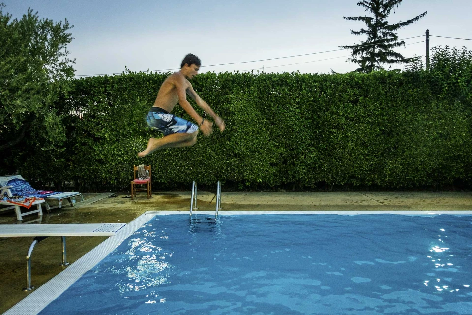 Teenage boy jumping into a backyard swimming pool, with diving board, pool ladder, and green hedge in the background on a summer day.