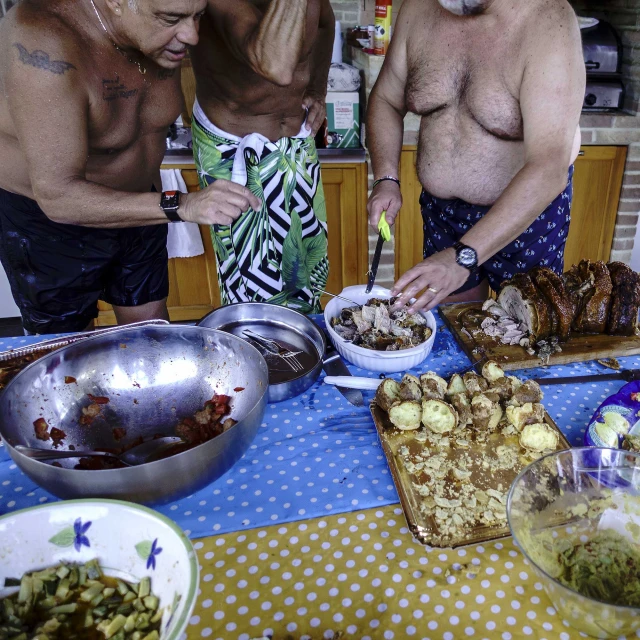 Three shirtless men preparing food at an outdoor table, featuring bowls of salad and potatoes, plus a roasted meat dish, on a sunny day.