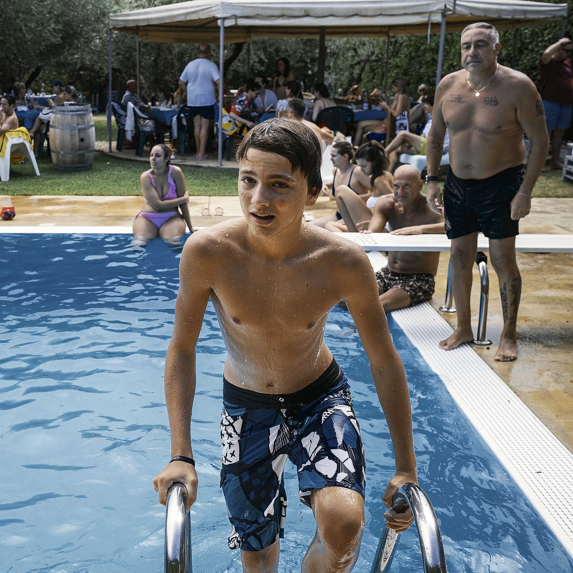 Teenage boy in patterned swim trunks climbing out of a blue swimming pool on a sunny day, with a group of people in the background.