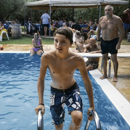 Teenage boy in patterned swim trunks climbing out of a blue swimming pool on a sunny day, with a group of people in the background.