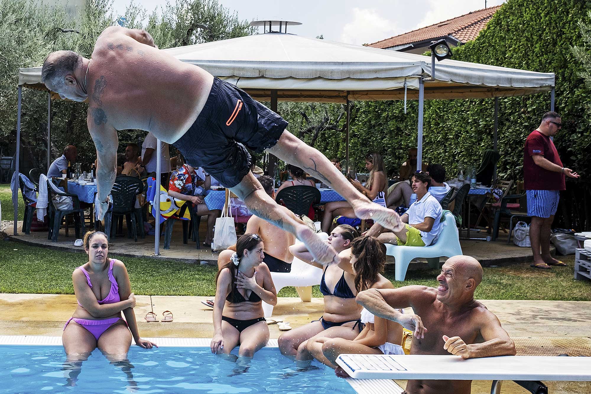 Adult male dives into outdoor pool at gathering. People sit poolside, while others dine at tables beneath a white canopy tent.