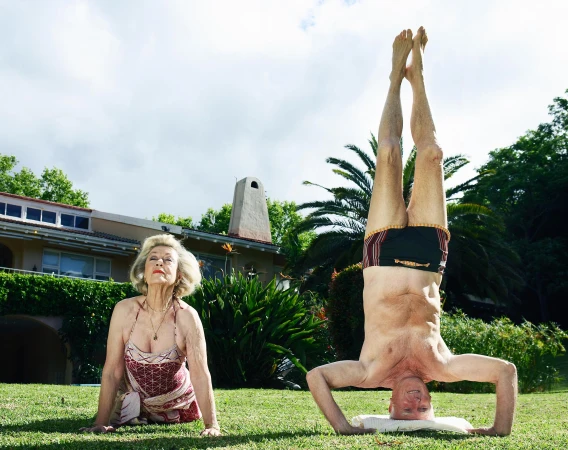 Mature adults practice yoga outdoors on a green lawn. One woman sits; a man is upside down in a headstand supported by a towel or pillow.