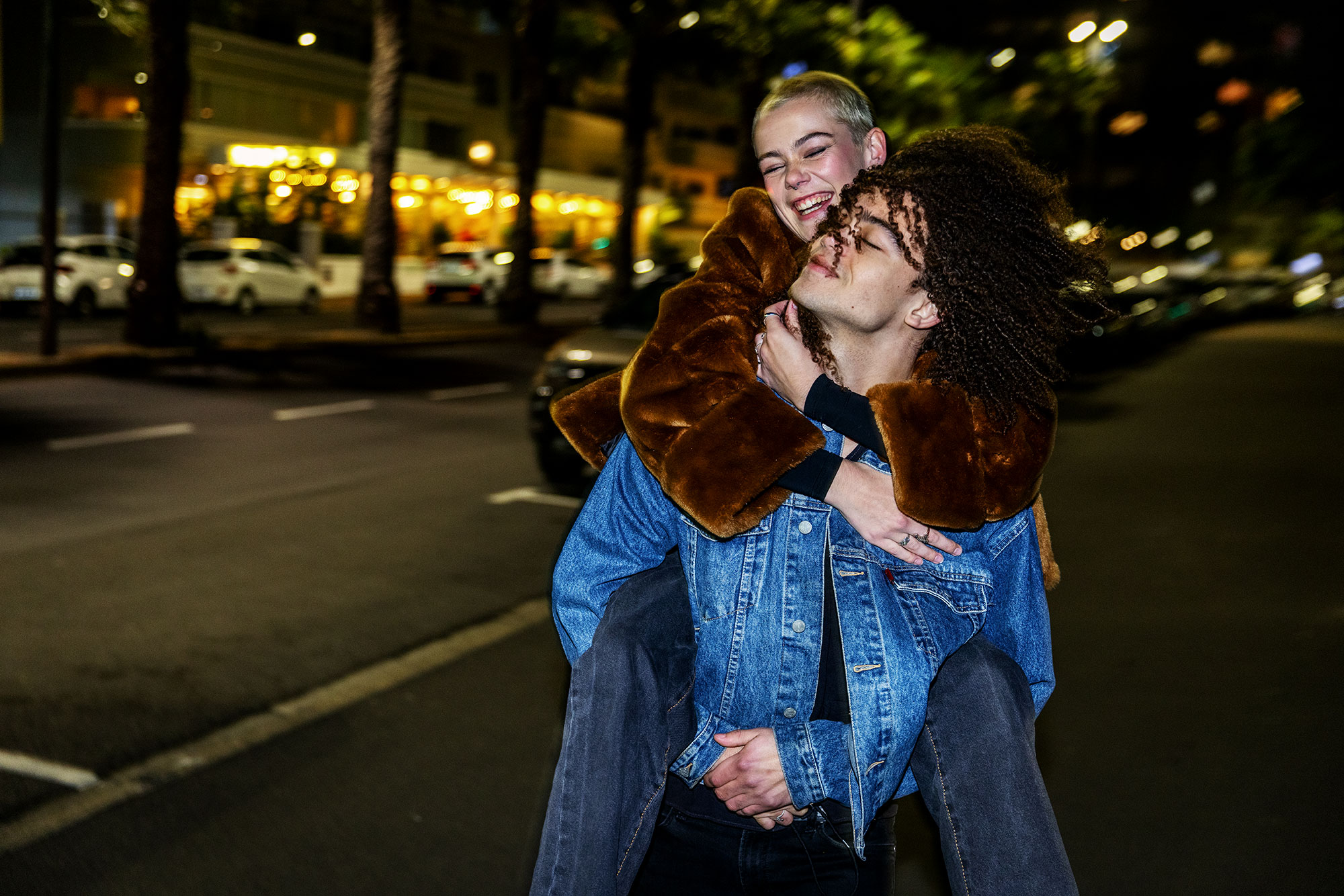 Happy young woman rides piggyback on a man on a city street at night with building lights in the background. Urban night portrait.