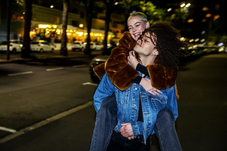 Happy young woman rides piggyback on a man on a city street at night with building lights in the background. Urban night portrait.