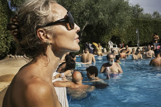 Close-up of an adult woman with sunglasses in a pool surrounded by a crowd of people; outdoor party scene with trees.