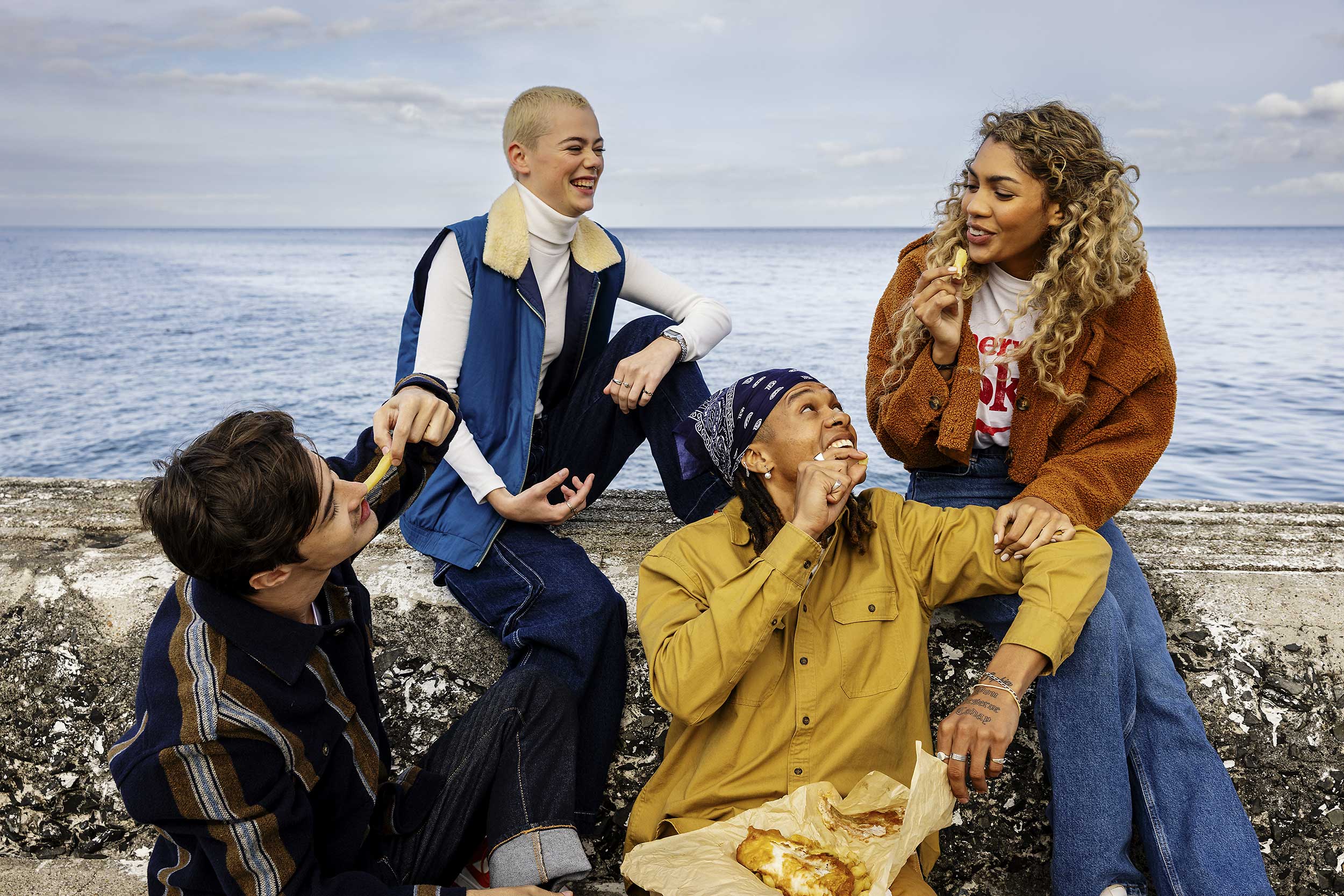 Four young adults sit together on a concrete ledge by the sea, eating snacks and laughing, dressed in casual clothing.