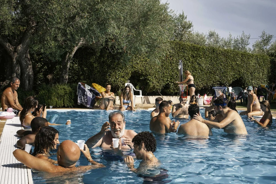 Outdoor pool party with a large group of people swimming, talking, and relaxing in an in-ground pool with trees and shrubs in the background.