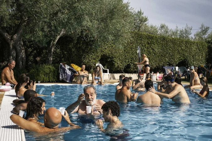 Outdoor pool party with a large group of people swimming, talking, and relaxing in an in-ground pool with trees and shrubs in the background.