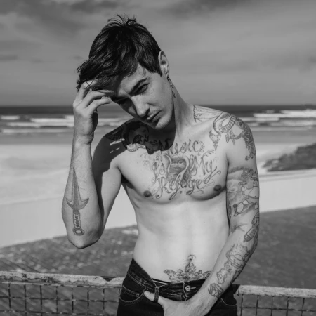 Black and white portrait of a shirtless tattooed young man posing outdoors with a coastal beach background.
