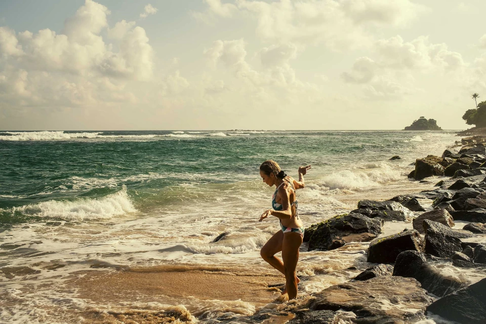 Woman in bikini walks on beach with ocean waves and rocky shoreline. Tropical paradise with cloudy skies, warm light and natural beauty.