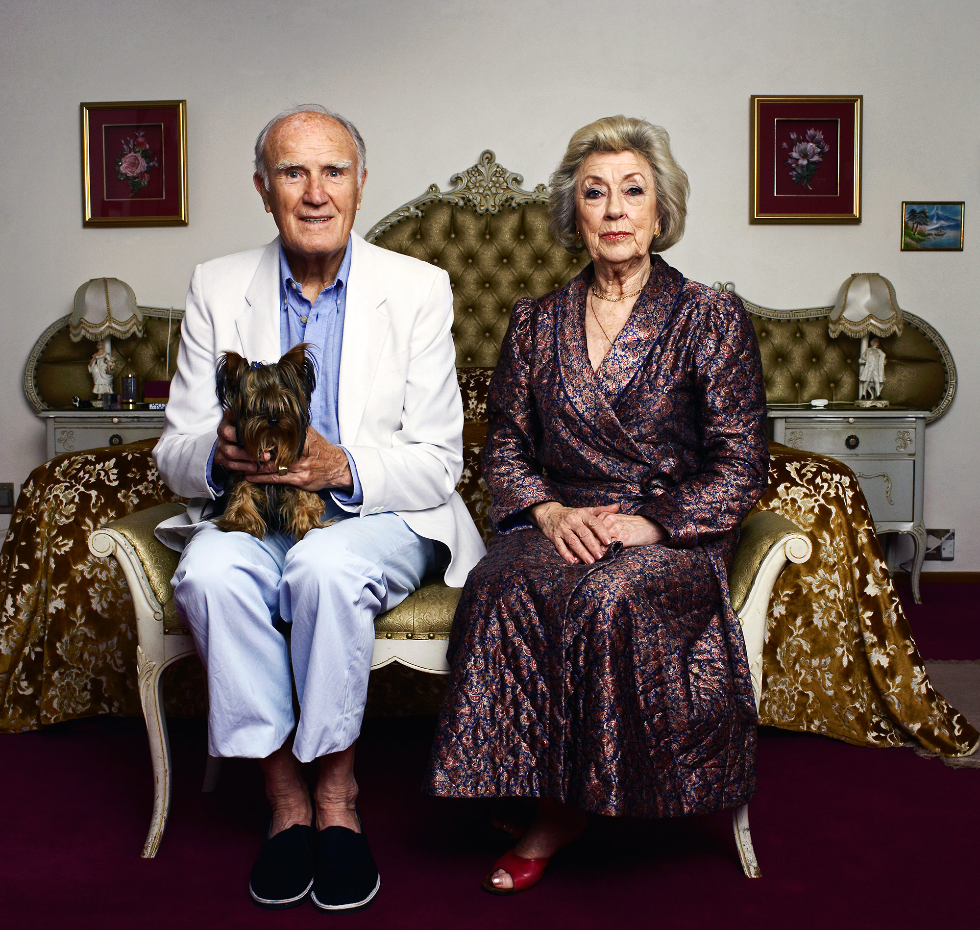 Portrait of an elderly couple seated indoors. The man holds a Yorkshire Terrier, and a large bed is behind them in the shot.