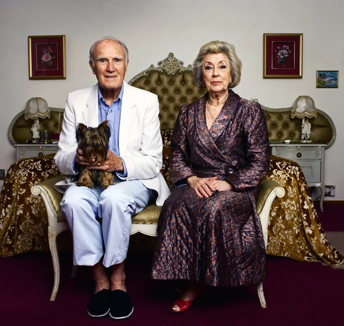 Portrait of an elderly couple seated indoors. The man holds a Yorkshire Terrier, and a large bed is behind them in the shot.