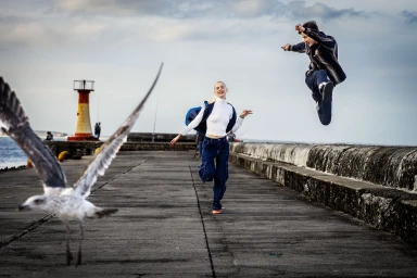 A seagull flying in the foreground while a woman runs and a man jumps on a concrete pier with a lighthouse in the distance