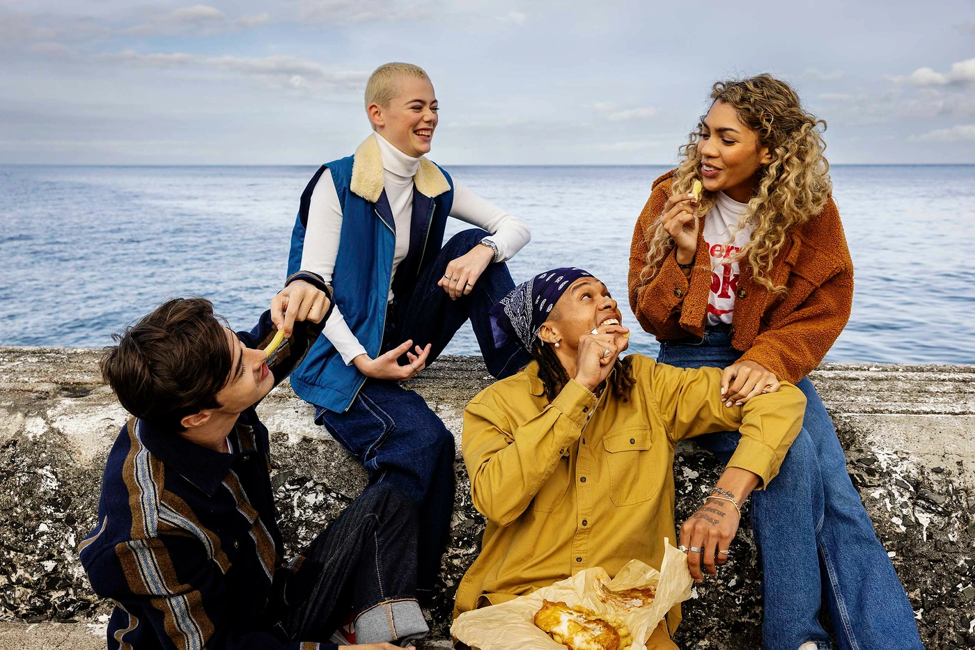 Four young friends laughing and sharing fish and chips while sitting on a concrete sea wall against an ocean background.
