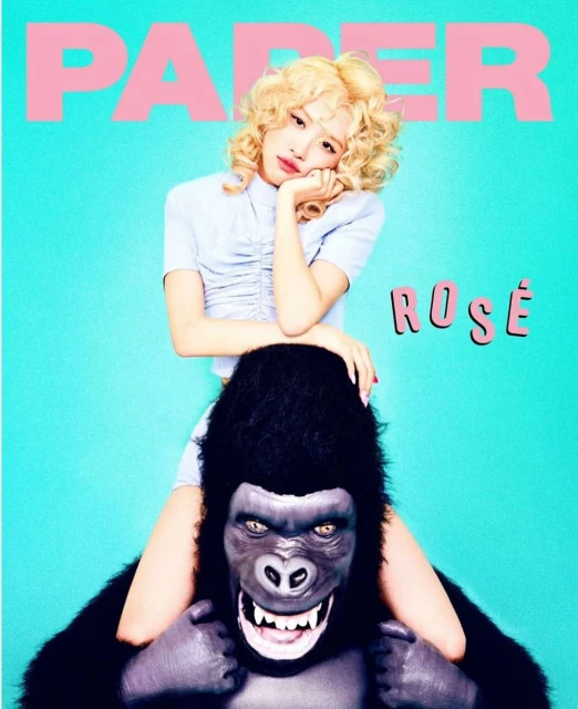 Young woman poses playfully on the shoulders of a gorilla mascot head in a studio shot with a bright turquoise backdrop and large pink text.