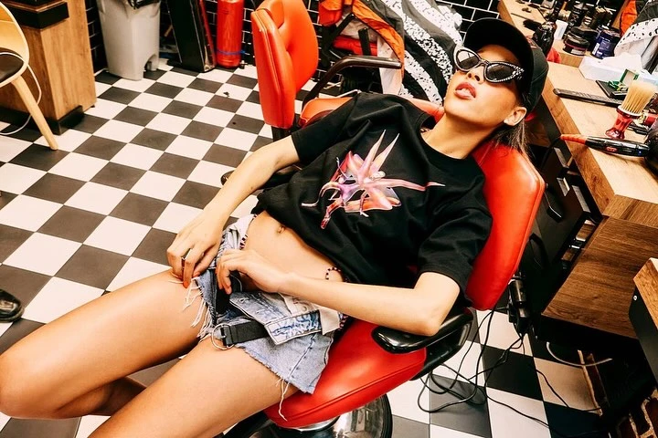 Woman in sunglasses and cap reclines in a red barber chair in a hair salon, posing with a black and white checkered floor.