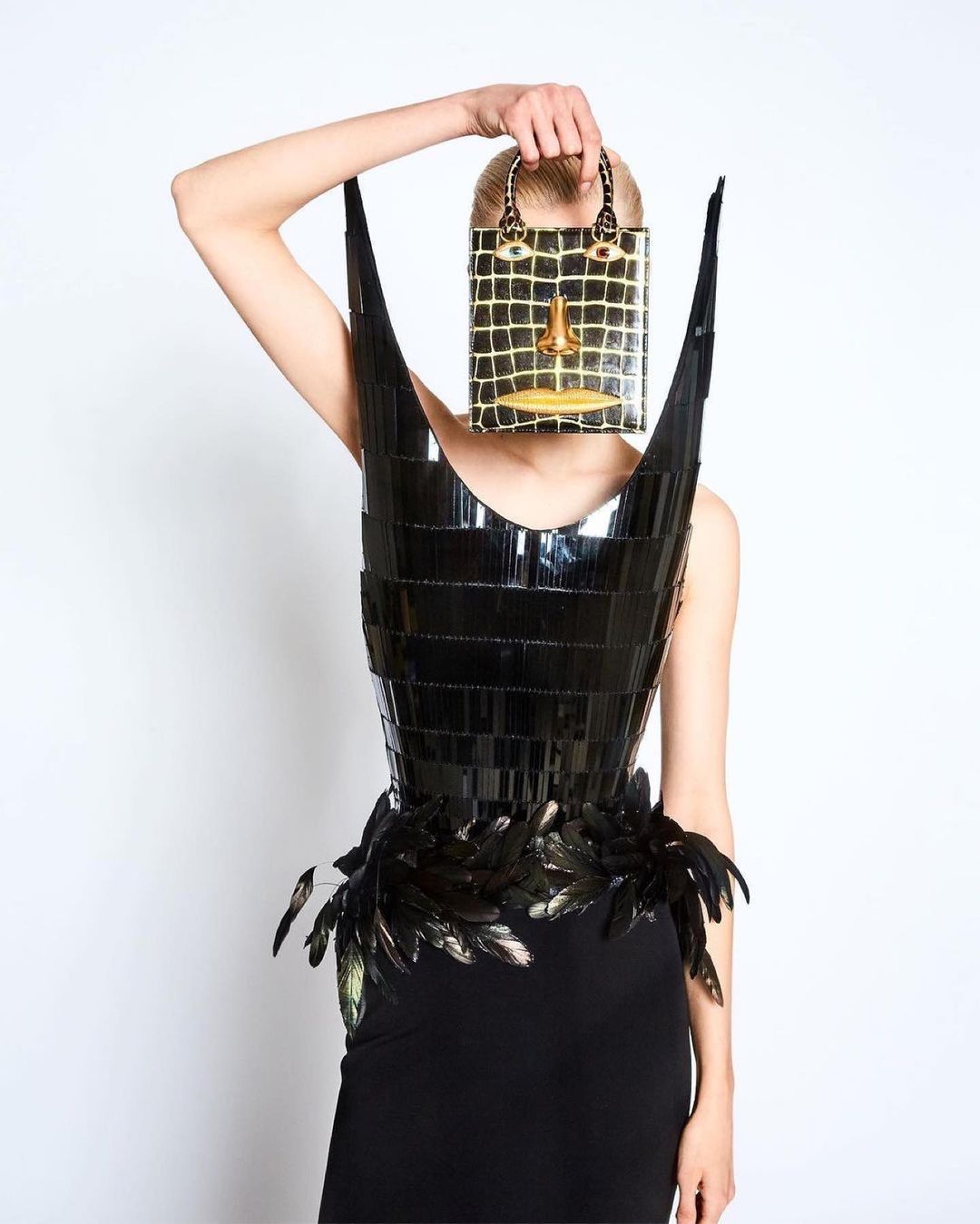 Fashionable woman wearing a glossy avant-garde dress with feather accents and holding a purse shaped like a face against a white backdrop.