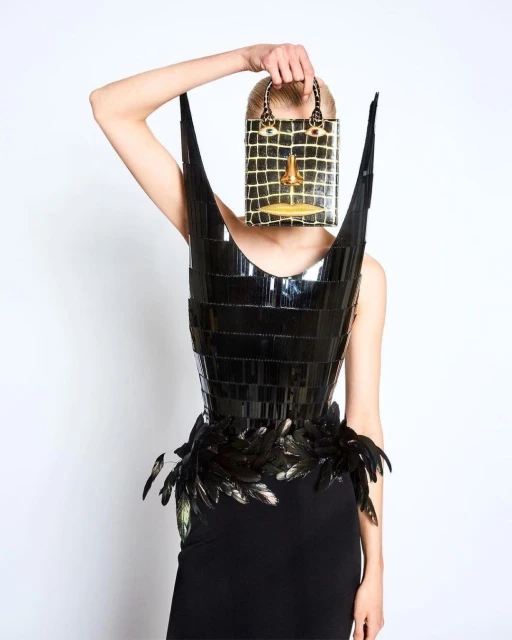 Fashionable woman wearing a glossy avant-garde dress with feather accents and holding a purse shaped like a face against a white backdrop.