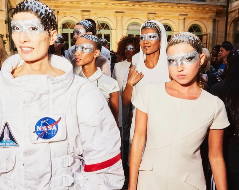 Fashion show featuring models wearing avant-garde silver makeup and outfits with one model wearing a white spacesuit with NASA logo.