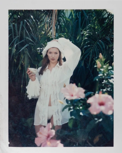 A woman in white swimwear and a hat poses holding a teacup amidst tropical foliage and hibiscus flowers in a dreamy outdoor setting.