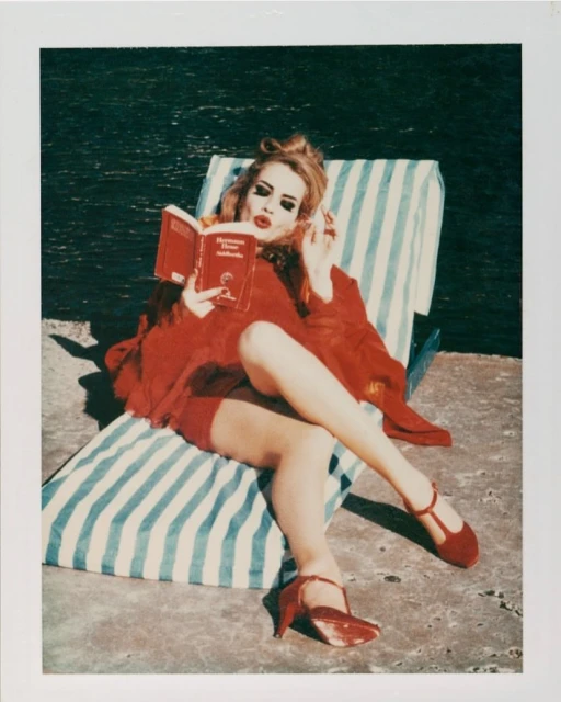 Woman in red dress reads book on striped lounge chair near water.  Adult female sits outside on sunny day with a book.