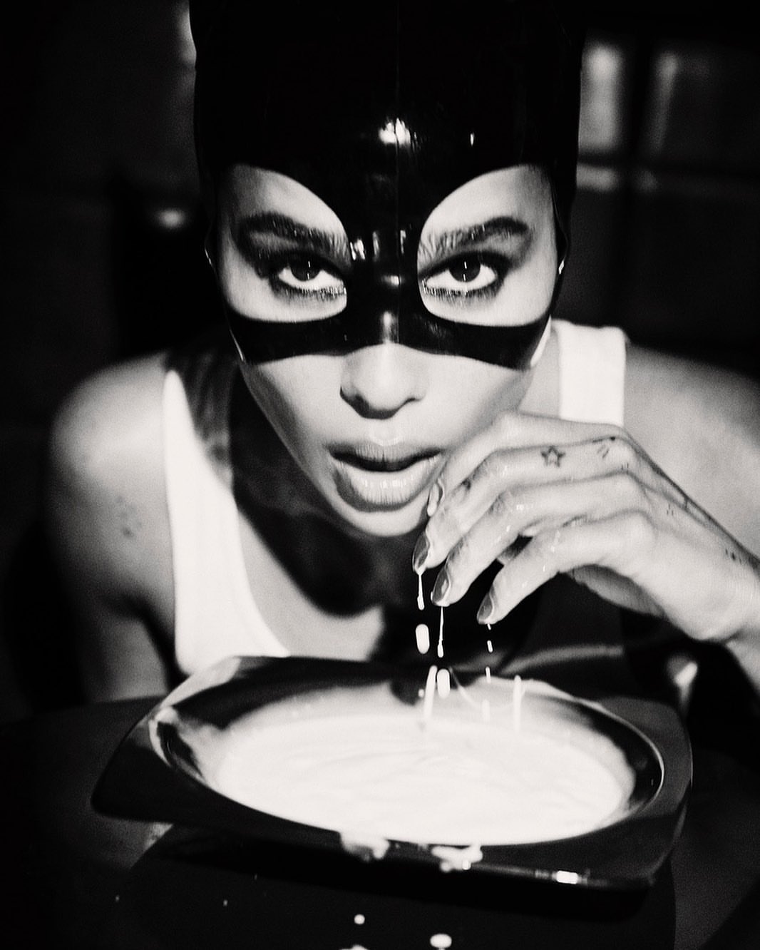Monochrome close-up of a woman in a mask, gazing at the camera while holding her hand over a bowl of white liquid.