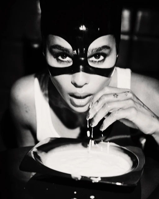 Monochrome close-up of a woman in a mask, gazing at the camera while holding her hand over a bowl of white liquid.