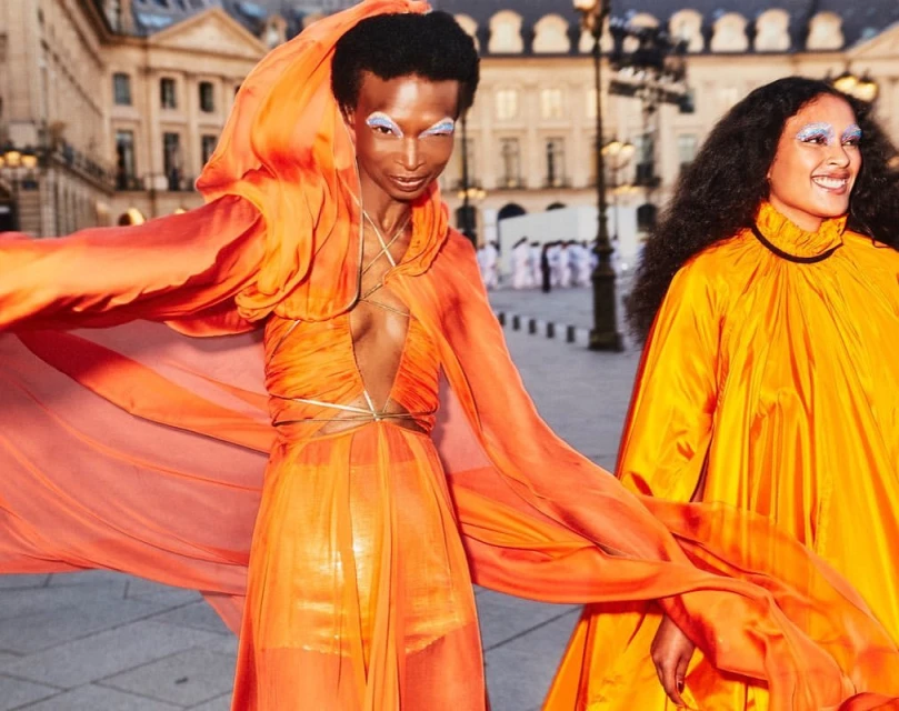 Two fashion models in orange outfits, posing in front of a building on a sunny day. One model has elaborate white eye makeup.