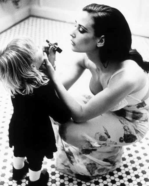 Black and white image of an adult woman applying makeup to a child. The woman is kneeling on a tiled floor, and the child is standing.