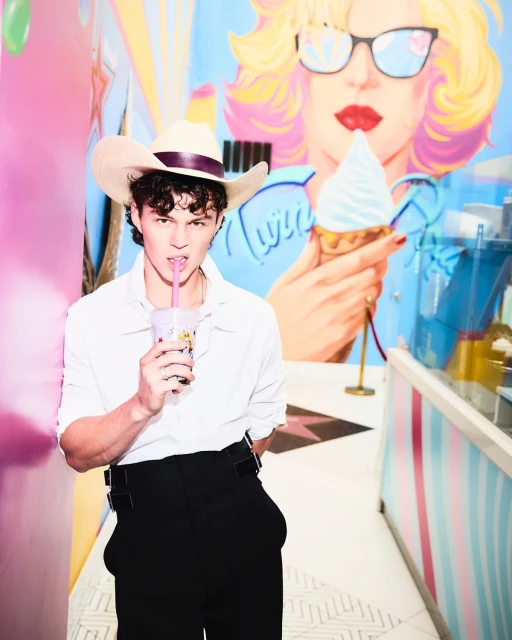 Young man in a white shirt and cowboy hat sips a drink with a straw, posing indoors in front of a mural, creating a colorful portrait.