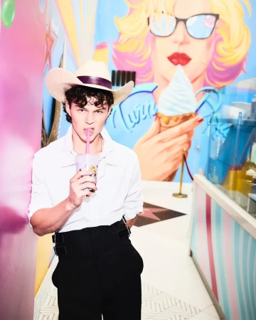 Young man in a white shirt and cowboy hat sips a drink with a straw, posing indoors in front of a mural, creating a colorful portrait.