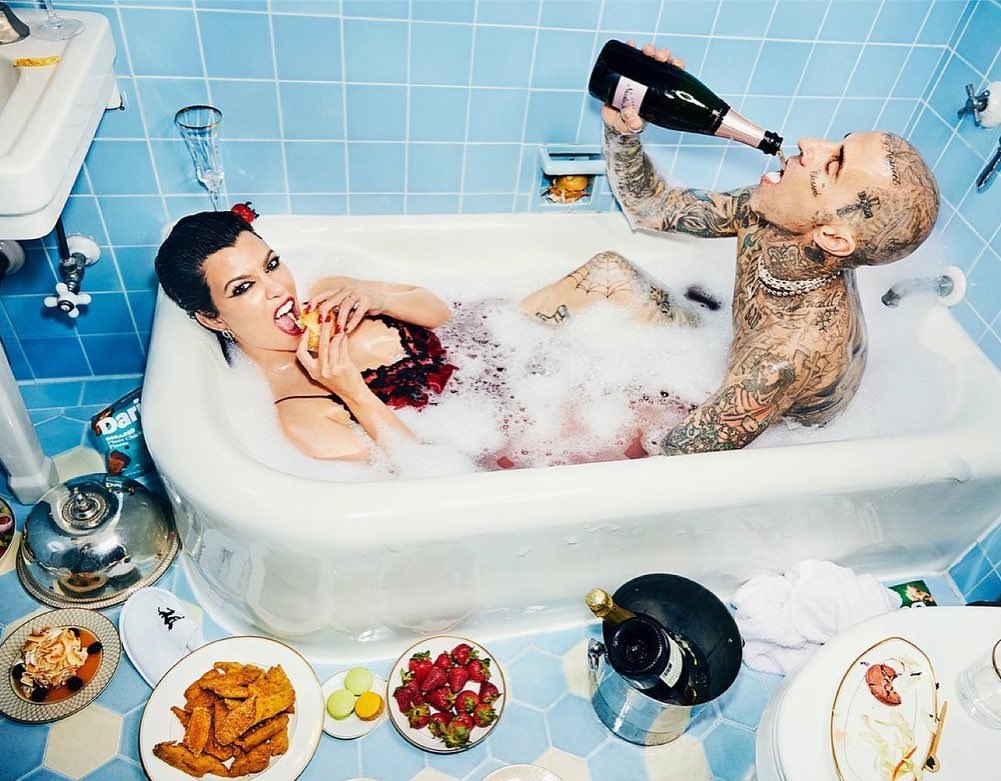 Kourtney Kardashian and Travis Barker relax in a bubble-filled bathtub, surrounded by food and champagne, under blue tiled walls.