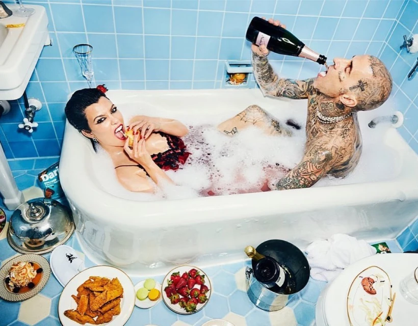 Kourtney Kardashian and Travis Barker relax in a bubble-filled bathtub, surrounded by food and champagne, under blue tiled walls.