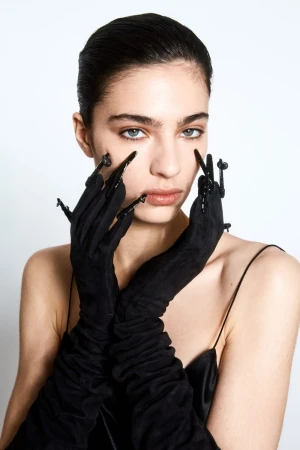 Close-up portrait of a woman with light skin, dark hair, and light-colored eyes wearing long, black gloves with black embellishments.