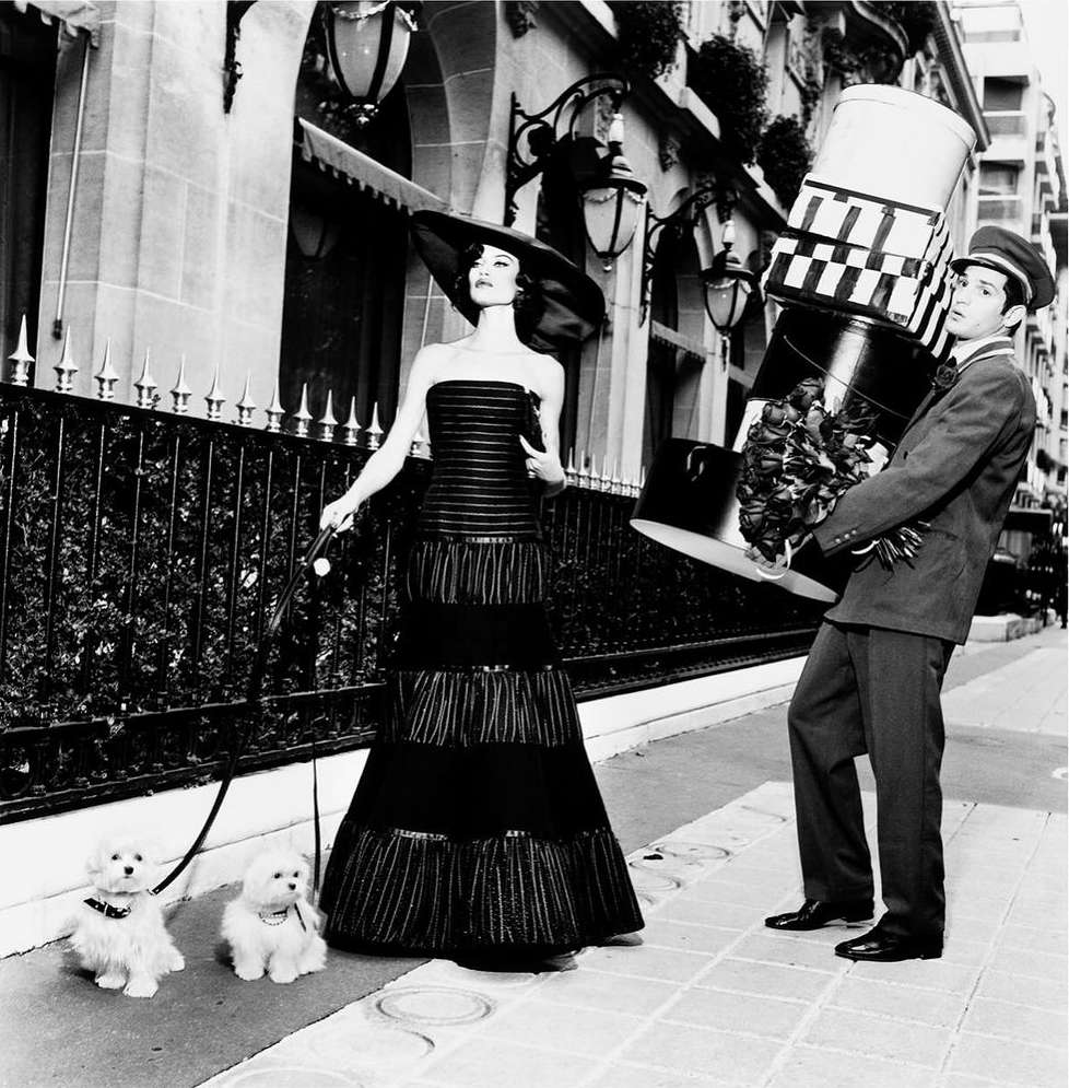 Black and white shot of a woman in a black dress walking two small white dogs with a man holding a stack of boxes.