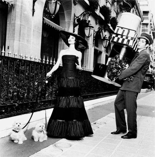 Black and white shot of a woman in a black dress walking two small white dogs with a man holding a stack of boxes.