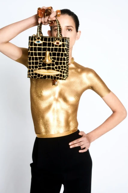 Fashionable woman with glittering gold body paint holds a purse that has a face design against a bright white background.