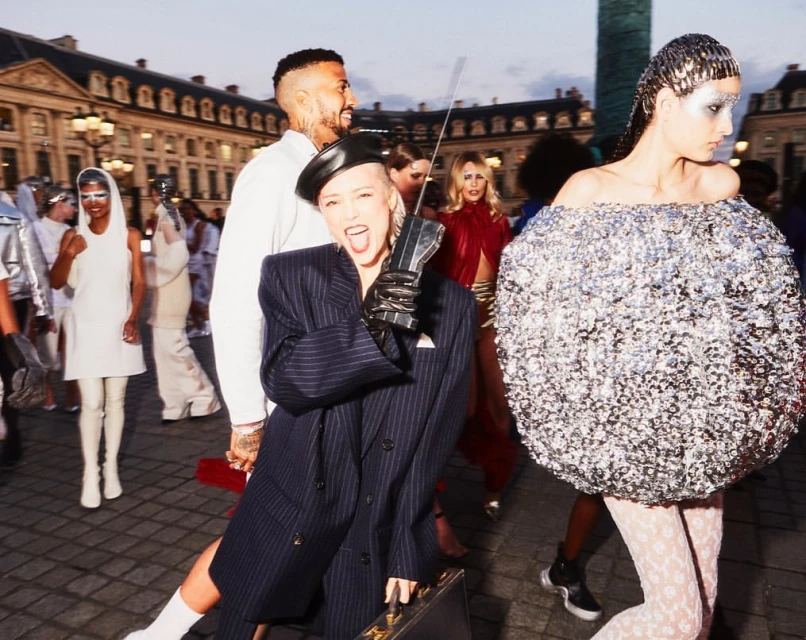 Outdoor fashion show scene in Paris featuring models in futuristic outfits and accessories, cobblestone ground, and classic architecture.