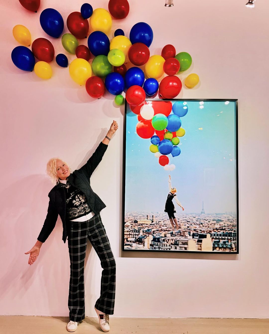 Woman in plaid pants posing with arm raised next to colorful balloons and framed photo of a woman with balloons over Paris.