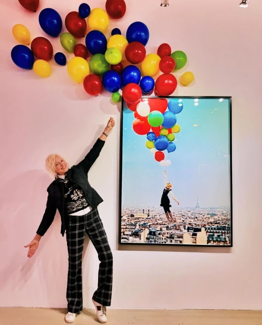 Woman in plaid pants posing with arm raised next to colorful balloons and framed photo of a woman with balloons over Paris.