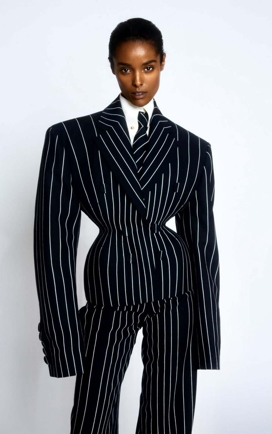 Fashionable African American woman in pinstripe suit with a narrow waist standing against a white backdrop in a studio setting.