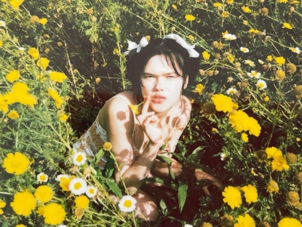Young adult female in a floral dress posing in a sunny field of yellow and white wildflowers with a white ribbon in her dark hair.