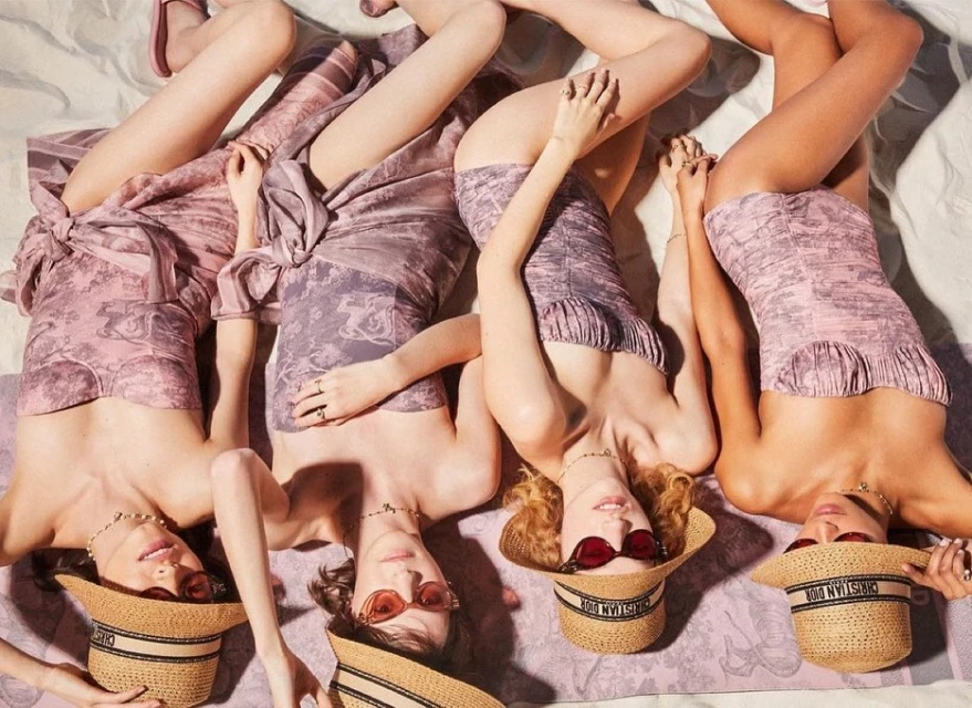 Four women lying on a patterned blanket, wearing matching swimsuits and hats in an eye-level shot in a beauty and fashion setting.