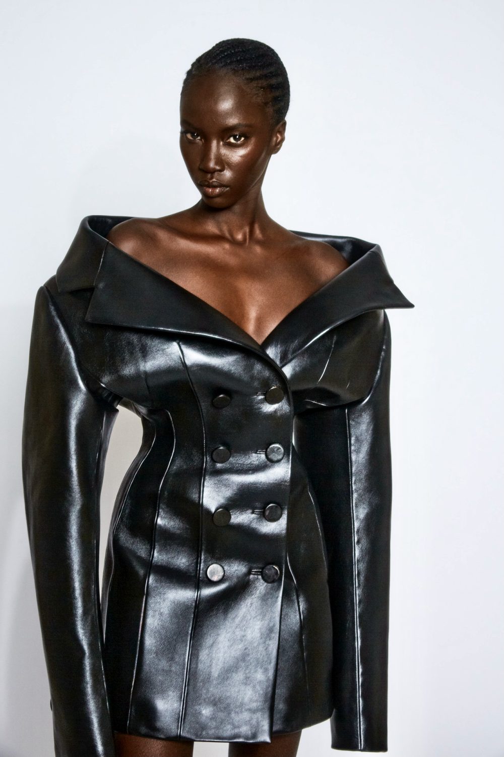 Fashionable dark-skinned woman poses in a stylish black, double-breasted leather dress with an oversized collar against a white backdrop.