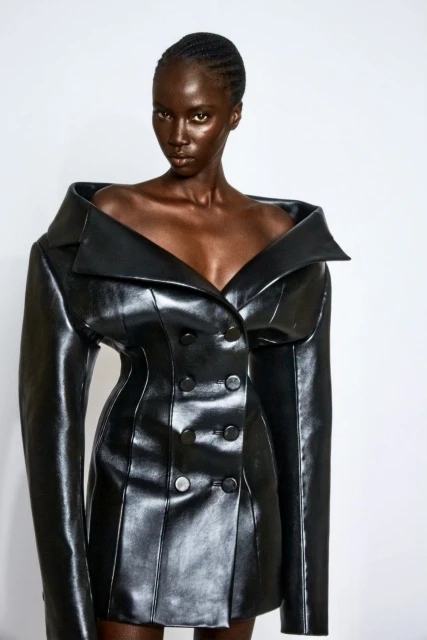 Fashionable dark-skinned woman poses in a stylish black, double-breasted leather dress with an oversized collar against a white backdrop.