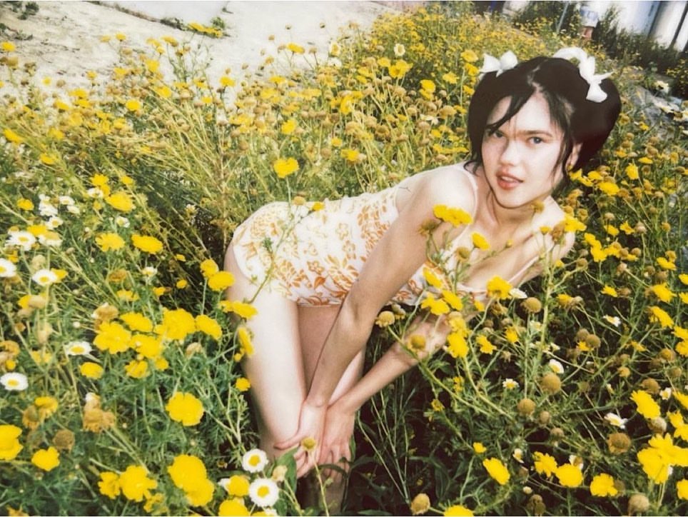 A young woman wearing a floral dress kneels among vibrant yellow wildflowers, with a soft, dreamy aesthetic and natural light.