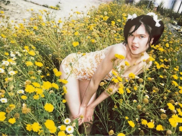A young woman wearing a floral dress kneels among vibrant yellow wildflowers, with a soft, dreamy aesthetic and natural light.