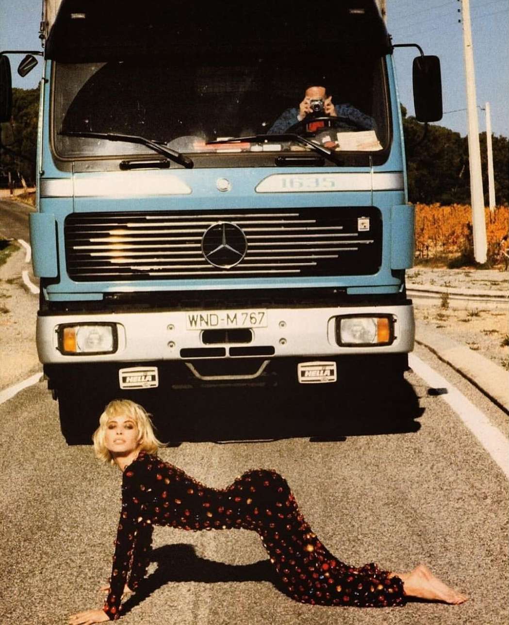 Fashion shot featuring a blond model in a sequined suit posing on an asphalt road with a blue Mercedes truck as a backdrop.