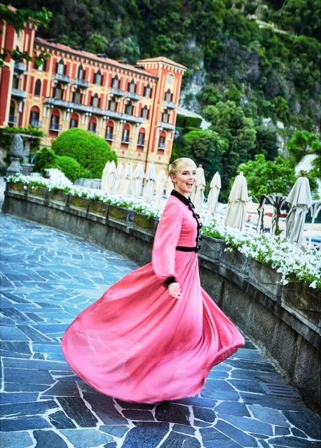 A woman twirls in a pink dress on a stone path near a grand hotel with white umbrellas and lush green trees in the background.